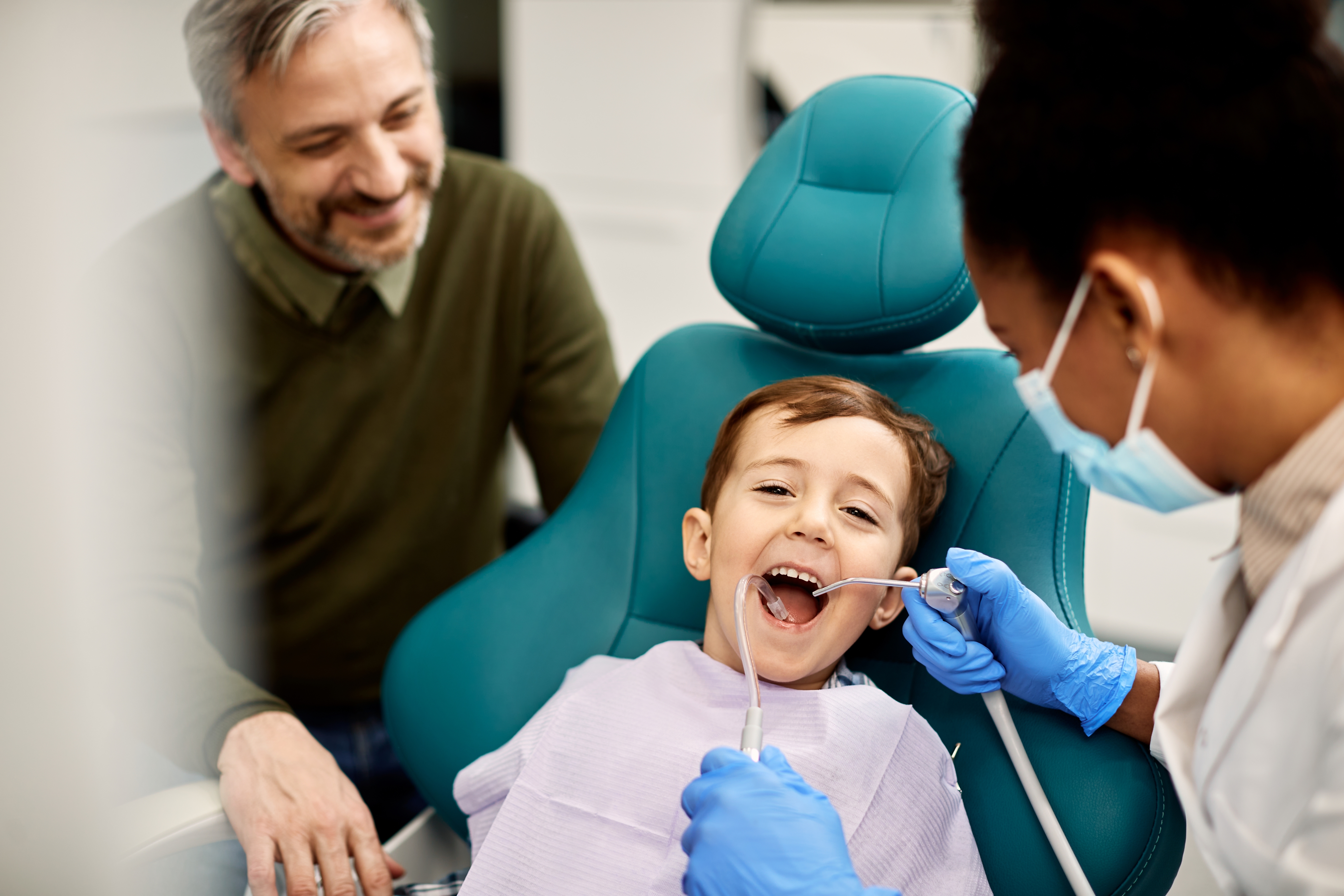 child in the dentist seat and father looking over him while dentist cleans teeth